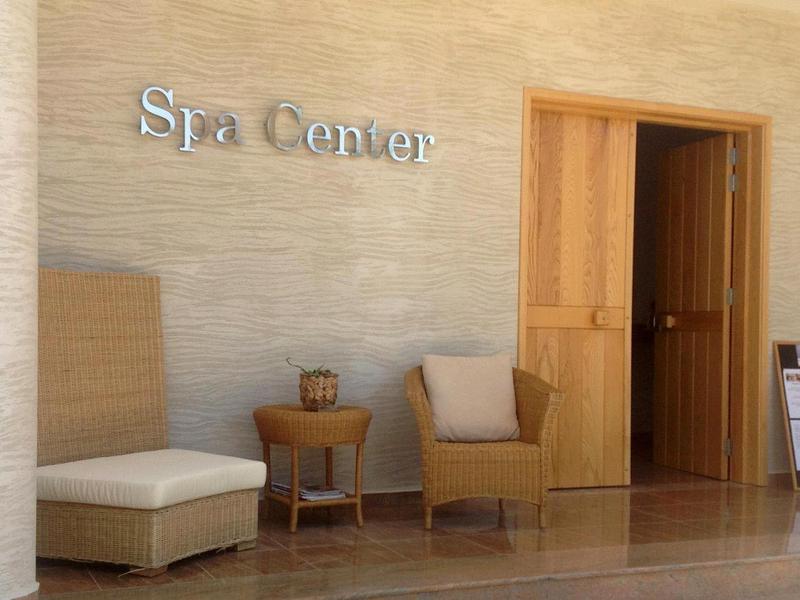 Wejście do spa z drewnianymi drzwiami, beżowym fotelem, podnóżkiem i małym stolikiem na drewnianej podłodze.