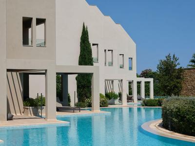 Piscina moderna de hotel con edificio blanco, agua azul y vegetación verde bajo un cielo soleado.