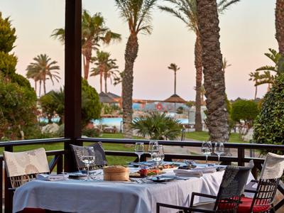 Terrassen-Tisch mit weißer Tischdecke, gedeckt mit Gläsern, Geschirr, Palmen und Pool im Hintergrund.