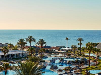 Blick auf einen Hotelpool mit Palmen und Meer im Hintergrund bei klarem Himmel.