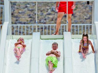 Drei Kinder rutschen nebeneinander eine breite weiße Wasserrutsche hinunter, darüber steht ein Mann.