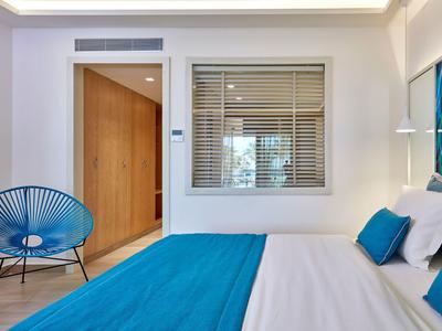 Schlafzimmer mit weißem Bett, blauem Bettüberwurf, blauer Stuhl und Holzdetails im Hintergrund.