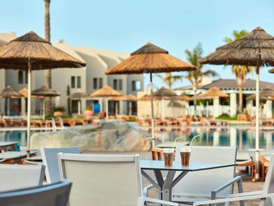 Área de piscina al aire libre con tumbonas, sombrillas y edificios del hotel al fondo bajo cielo despejado.