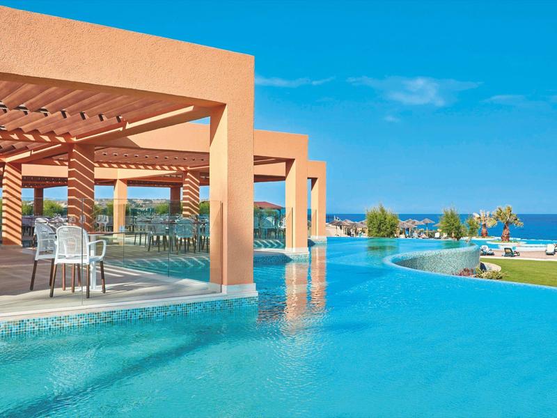 Moderner Pool mit türkisfarbenem Wasser, orangefarbenen Pavillons und Blick aufs Meer unter blauem Himmel.