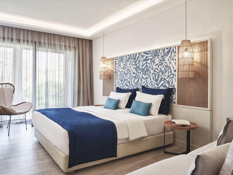 Modernes Schlafzimmer mit großem Bett, blauer Bettdecke, zwei Nachttischen und Sessel am Fenster.