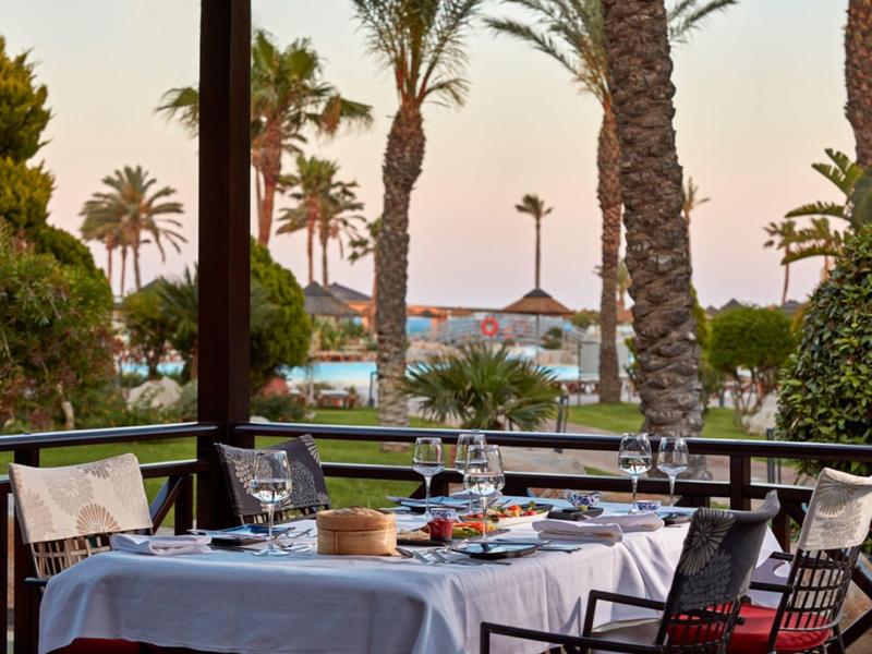 Terrassen-Tisch mit weißer Tischdecke, gedeckt mit Gläsern, Geschirr, Palmen und Pool im Hintergrund.