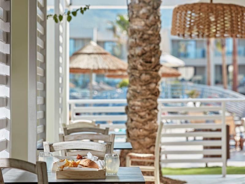 Hell eingerichtete Terrasse mit weißen Stühlen, Tisch und Blick auf Palmen und Sonnenschirme.