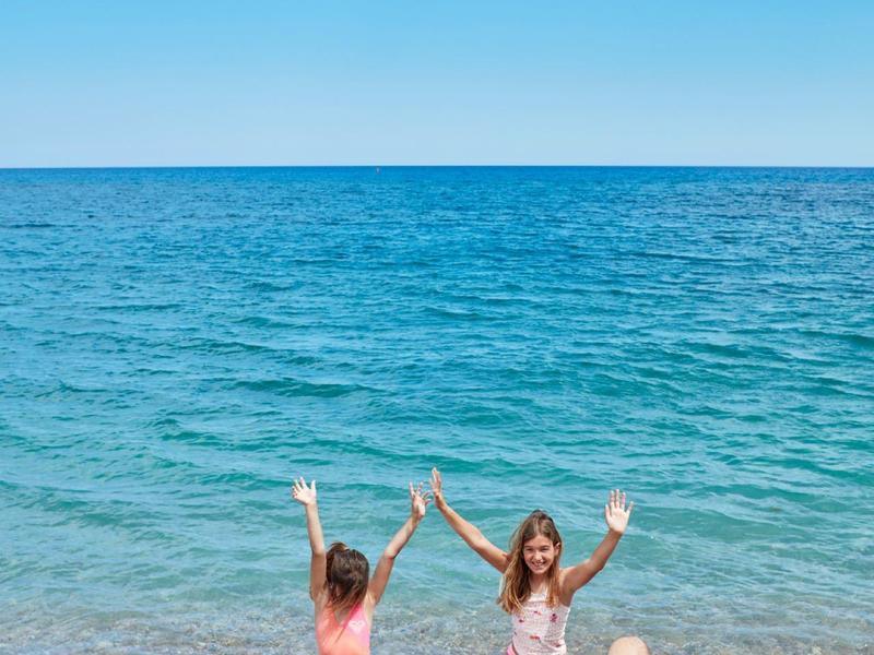 Vier Personen am Strand, zwei sitzen auf Kies, zwei stehen im Wasser, blauer Himmel und Meer im Hintergrund.