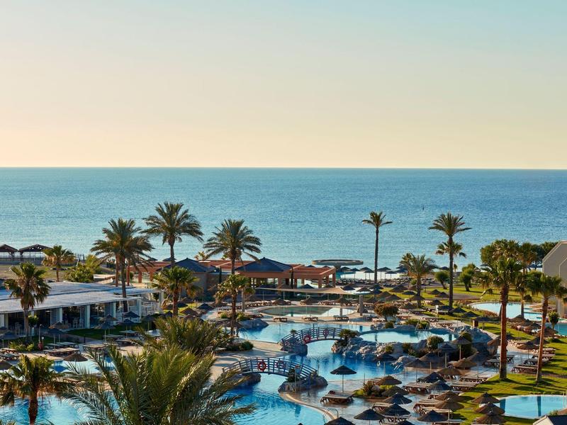 Blick auf einen Hotelpool mit Palmen und Meer im Hintergrund bei klarem Himmel.