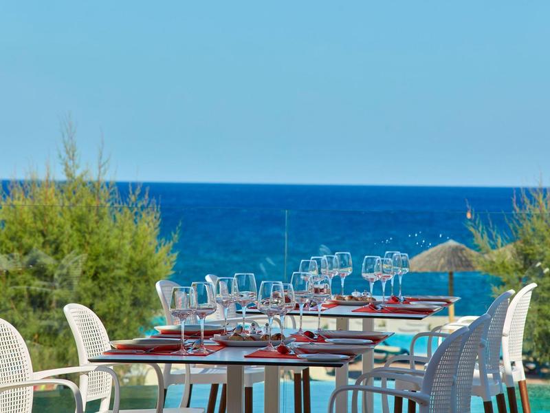 Terrassentische mit weißen Stühlen, gedeckt mit Gläsern und Besteck, vor blauem Meer und grünem Buschwerk.