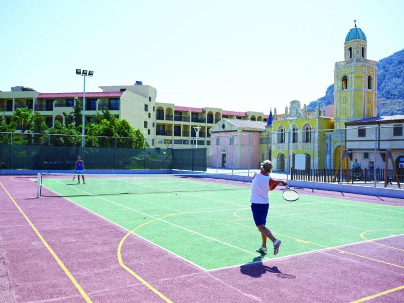 Tennisspieler auf farbigem Tennisplatz, Gebäudekomplex mit Turm im Hintergrund bei hellem Tageslicht.