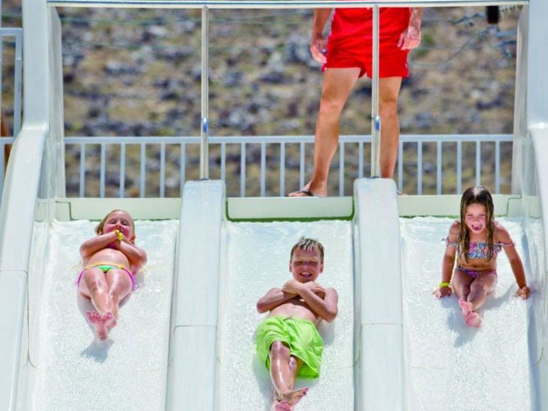 Drei Kinder rutschen nebeneinander eine breite weiße Wasserrutsche hinunter, darüber steht ein Mann.