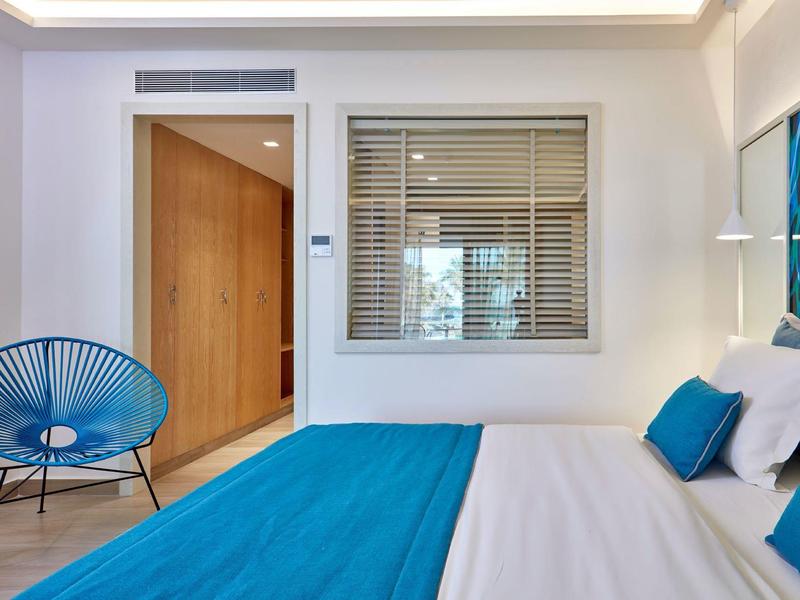 Schlafzimmer mit weißem Bett, blauem Bettüberwurf, blauer Stuhl und Holzdetails im Hintergrund.