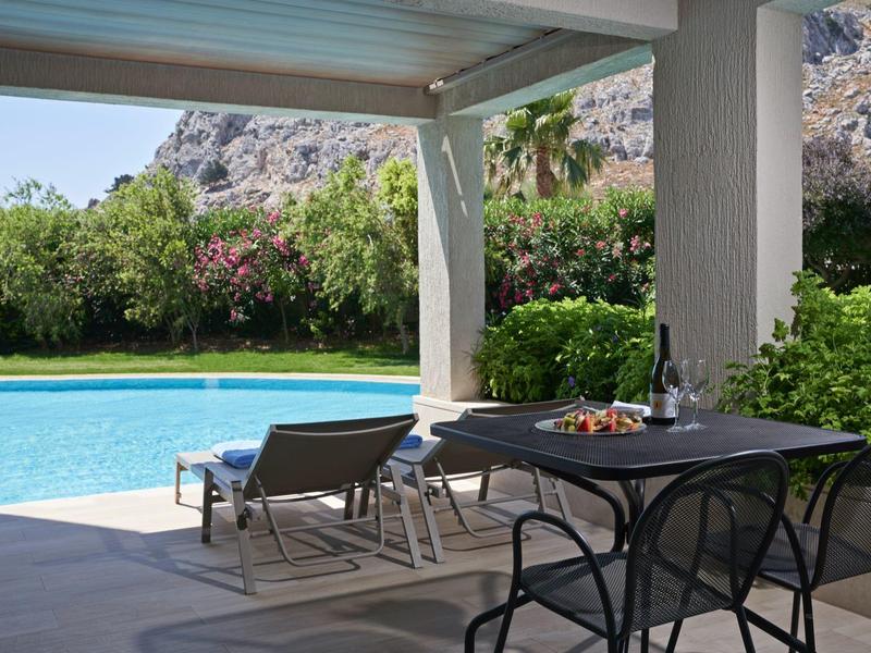 Überdachte Terrasse mit schwarzem Tisch und Stühlen, Blick auf Pool, Liegestühle, grüne Pflanzen und Berge.