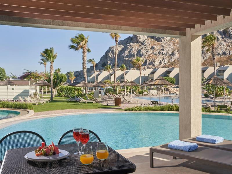 Blick auf Pool mit Liegen, grüner Rasen und Palmen vor felsigem Hügel unter blauem Himmel.