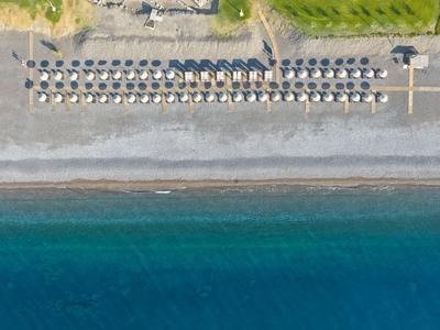 Widok z lotu ptaka na plażę z rzędami parasoli, żwirowym brzegiem i niebieskim morzem.