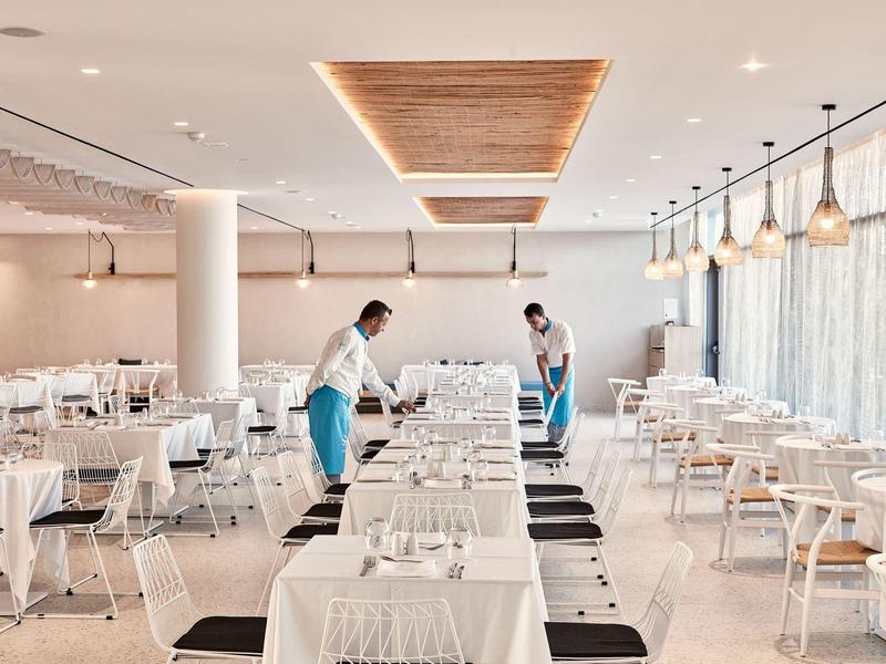 Großes, helles Restaurant mit weißen Tischen, Holzstühlen und zwei Kellnern, die Tische vorbereiten.