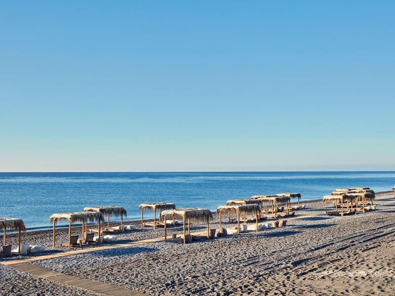 Strand mit Kies, mehreren Sonnenschirmen und Liegen vor ruhigem blauem Meer unter klarem Himmel.