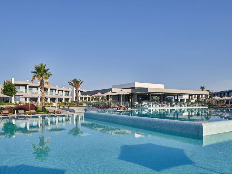 Großer Pool vor modernem Hotel mit Palmen und klarem blauem Himmel im Hintergrund.