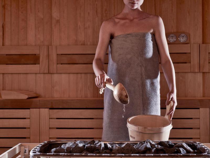 Person in Sauna mit grauem Handtuch gießt Wasser auf heiße Steine für Dampf.