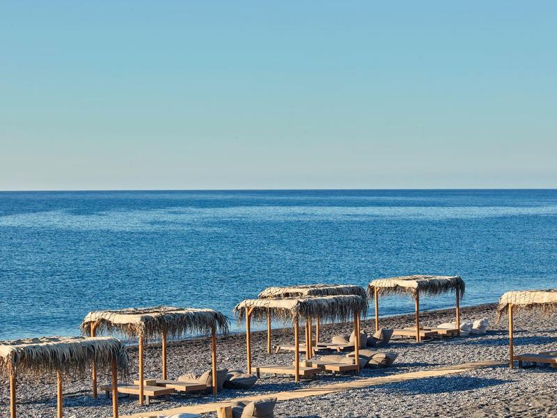 Strand mit mehreren leeren Strohdächern und ruhigem blauem Meer im Hintergrund unter klarem Himmel.