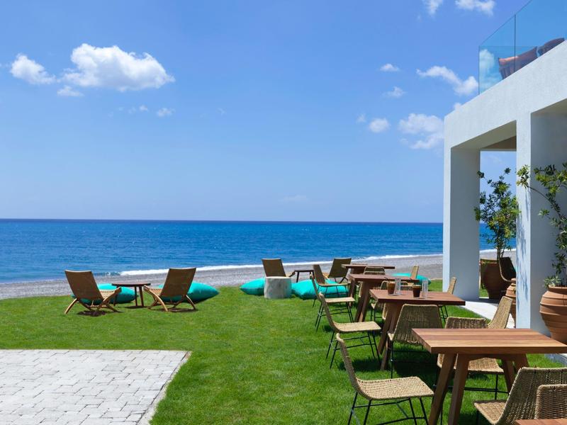 Strand mit blauem Himmel, Meer, Liegestühlen auf Gras und Tischen neben weißem Gebäude.