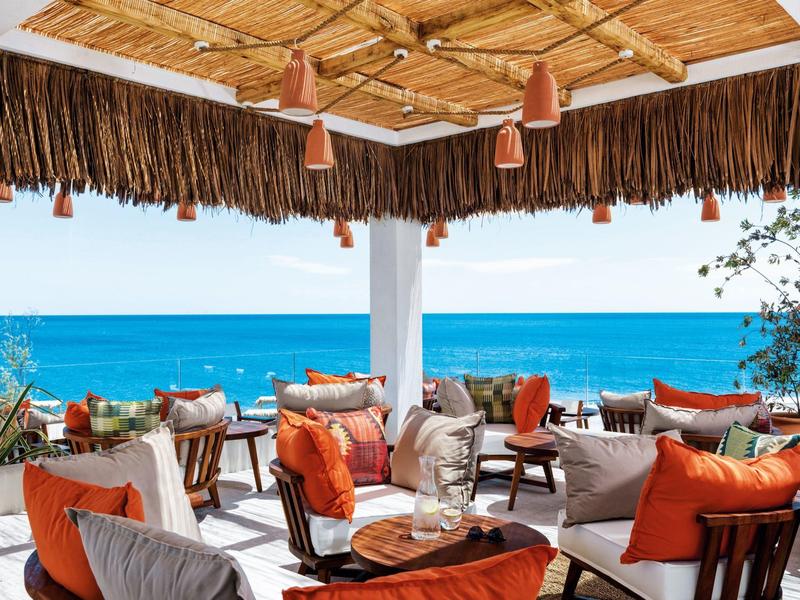 Terrassenbereich mit gemütlichen Sitzmöbeln und orangen Kissen, Blick auf blaues Meer und Himmel.