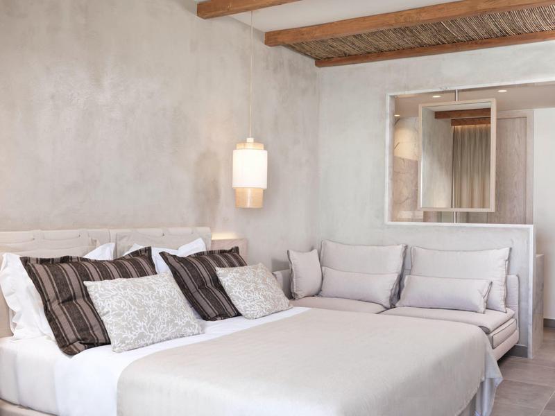 Helles Schlafzimmer mit großem Bett, mehreren Kissen und grauer Sofaecke vor Betonwand.