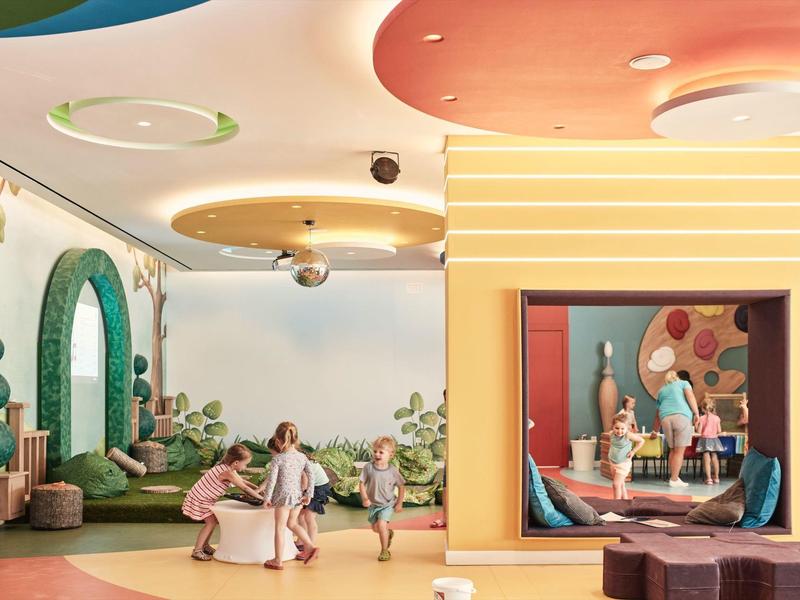 Bunte Kinderspielecke mit Sitzkissen, Kletterwand, großen Spiegeln und Kindern beim Spielen.