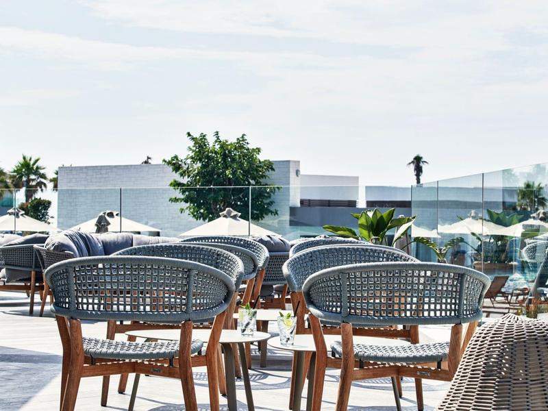 Leere Korbstühle um einen Tisch auf einer Terrasse mit Blick auf einen Pool und moderne Gebäude.