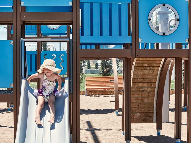 Kind mit Hut rutscht auf weißer Rutsche in blau-braunem Klettergerüst auf Sandboden.