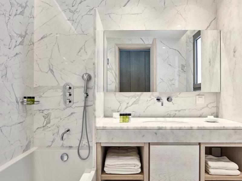 Weißer Marmorbad mit Badewanne, Handbrause, großem Spiegel und Waschtisch mit Handtüchern.