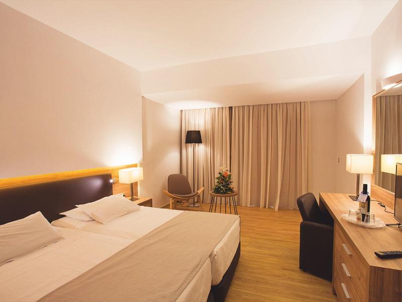 Zimmer mit Doppelbett, großen Vorhängen, Schreibtisch und warmem Licht in Holzoptik.