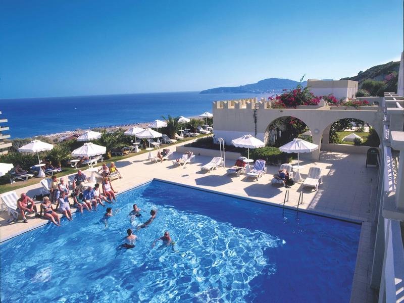 Ein blaues Schwimmbecken mit Menschen, umgeben von Liegestühlen und weißen Gebäuden am Meer.