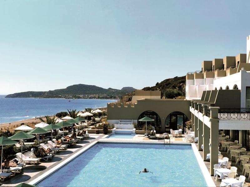 Blick auf Poolanlage mit Liegen, Sonnenschirmen und Gebäuden am Meer bei klarem Himmel.