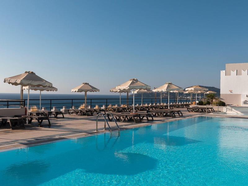 Großer blauer Pool mit Liegestühlen und Sonnenschirmen, dahinter klarer Himmel und Meer.