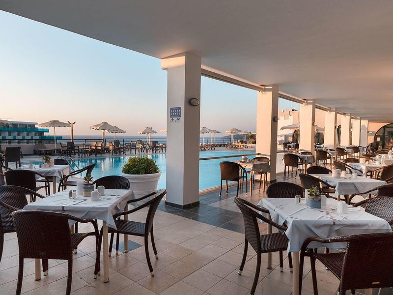 Terrassenrestaurant mit weißen Tischen, schwarzen Stühlen und Blick auf Pool und Meer bei Sonnenuntergang.
