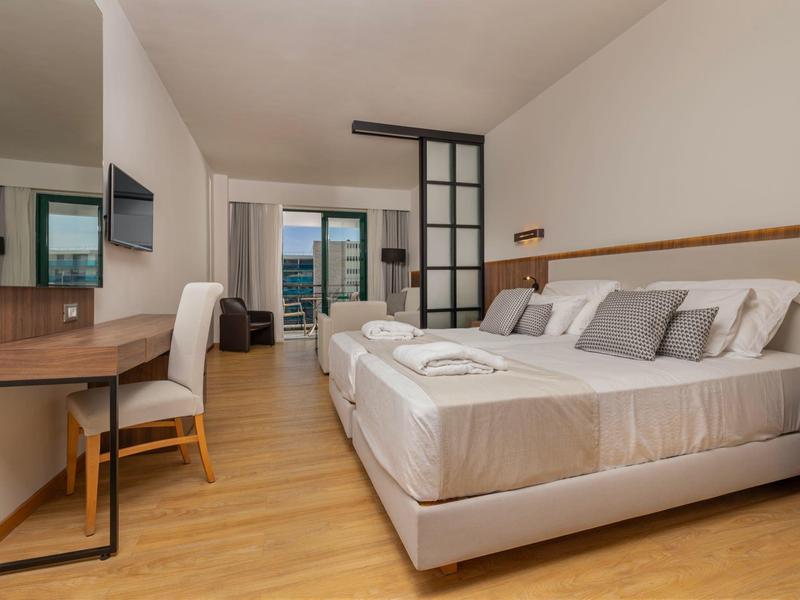Modernes Hotelzimmer mit zwei Betten, Schreibtisch, TV und Balkon mit Meerblick.