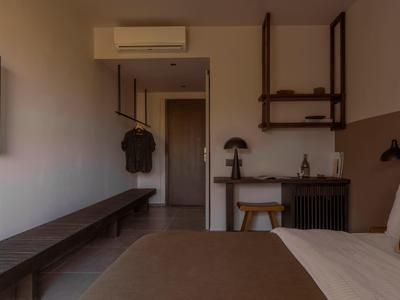 Kleines Zimmer mit braunem Bett, langer Bank, Hocker, Schreibtisch und Klimaanlage an der Wand.