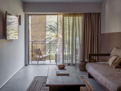 Gemütliches Wohnzimmer mit Sofa, Teppich und Couchtisch, Blick auf Balkon mit Stuhl und Palmen.