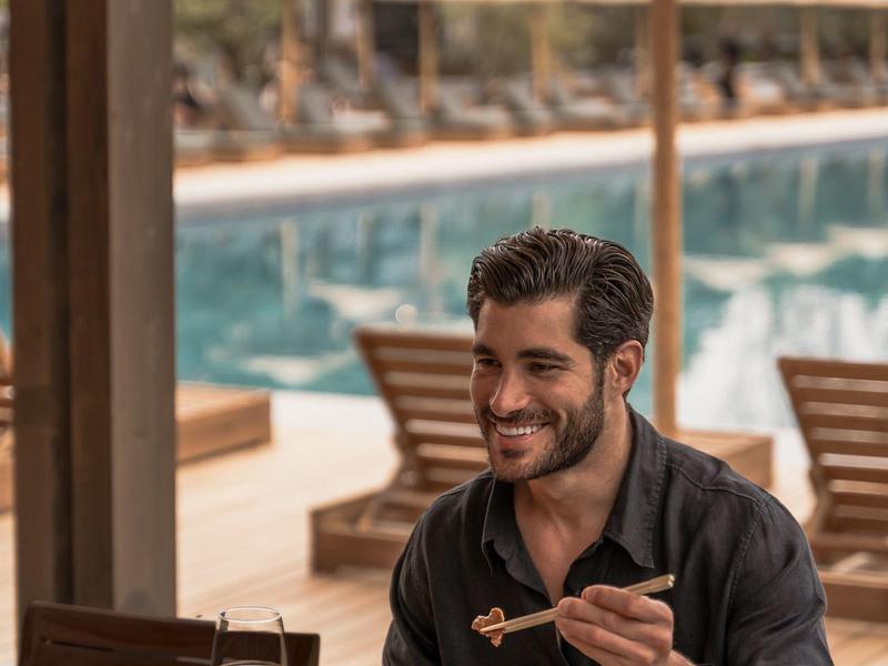 Uomo seduto vicino alla piscina che mangia con le bacchette a un tavolo all'aperto.