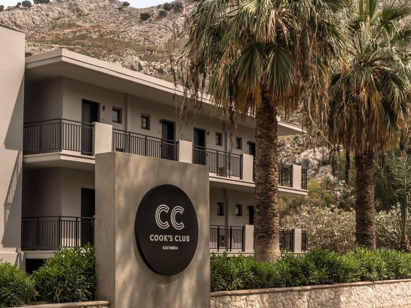 Complesso alberghiero moderno con palme e montagne sullo sfondo sotto un cielo limpido.