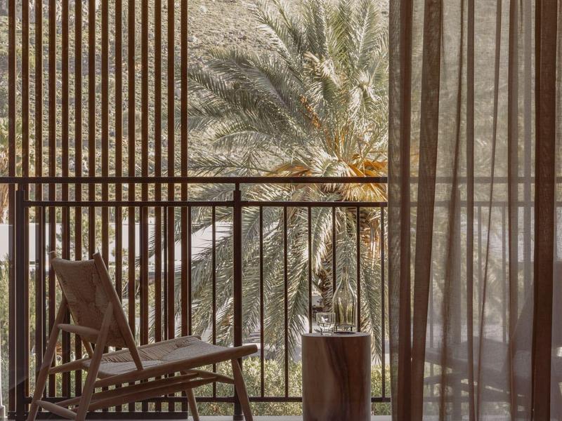 Balcone con sedia, tavolo e tende, vista su palme e sfondo roccioso.