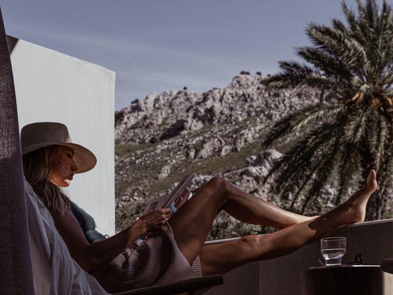 Donna rilassata su balcone con montagne e palma sullo sfondo al tramonto.