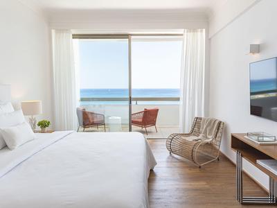 Helles Hotelzimmer mit Doppelbett, Stühlen und Balkon mit Meerblick.