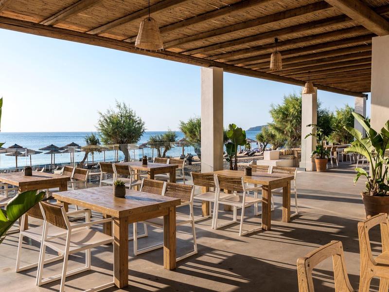 Terrazza coperta con tavoli e sedie in legno con vista mare e ombrelloni sulla spiaggia.