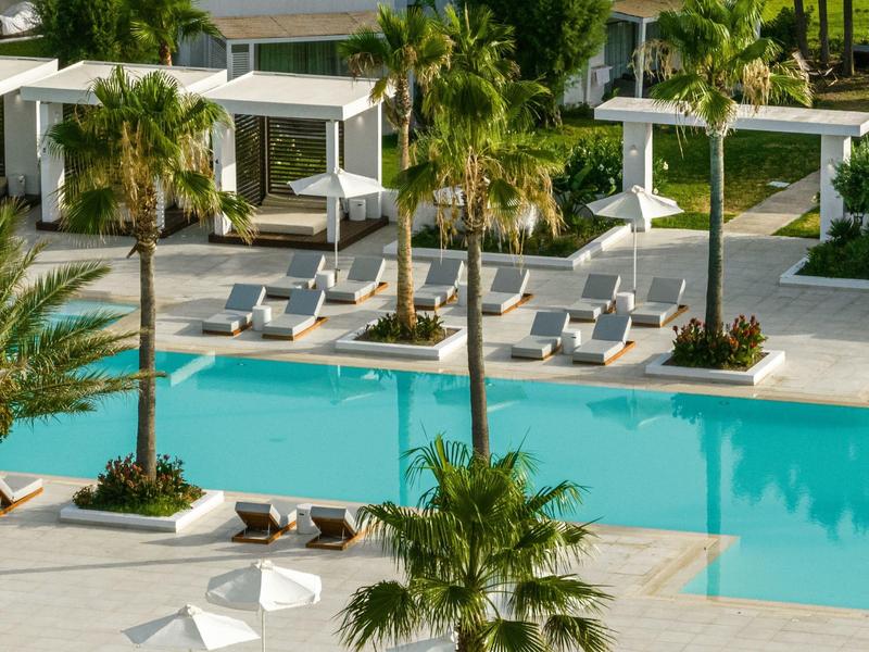 Area piscina moderna in hotel con palme, lettini e tende bianche.