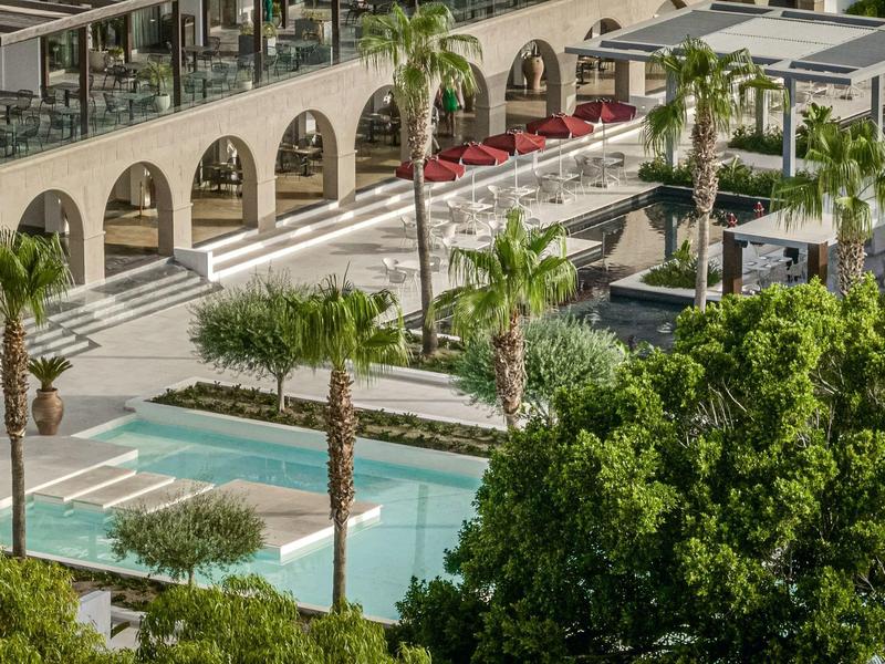 Terrazza dell'hotel con palme, piscina e ombrelloni rossi circondati da vegetazione.