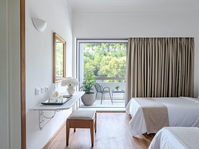 Camera d'hotel luminosa con balcone, tavolo, sedie e vista su alberi verdi.