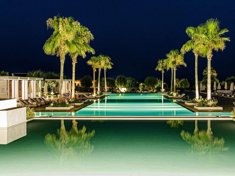 Piscina esterna illuminata con palme e riflessi chiari di notte in un hotel.